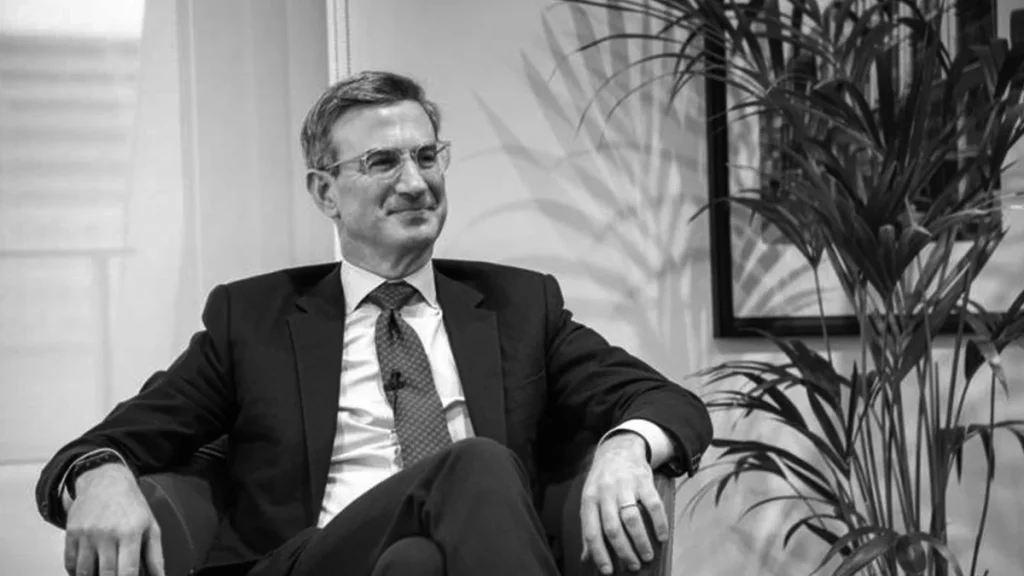 Peter Orszag sitting in a chair wearing a suit and glasses, showcasing his neat side-part haircut often referred to as the “Power Cut.”